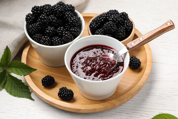 Fototapeta na wymiar Fresh ripe blackberries, tasty jam and leaves on white wooden table, closeup