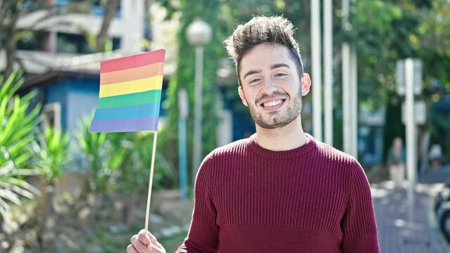 Young hispanic man smiling confident holding rainbow flag at park