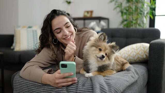 Young hispanic woman with dog using smartphone lying on sofa at home