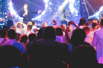 Crowded concert hall arena with scene stage lights with musicians band on a stage at the venue, rock show performance, with concert-goers attendees, audience on dance floor during concert festival