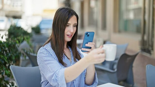 Young beautiful hispanic woman make photo to cup of coffee by smartphone smiling at coffee shop terrace