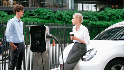 Progressive businessman and businesswoman leaning on electric car connected to charging station...