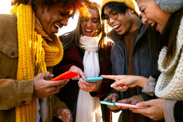 Cheerful multiracial friends looking at mobile phone together on winter day. Young woman pointing at device screen.