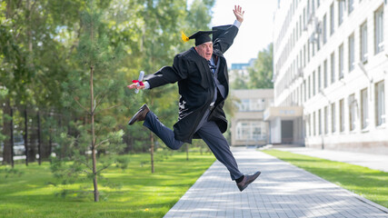 Old happy man in graduation gown jumping outdoors and holding diploma.