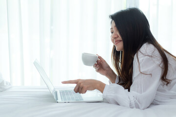 Asian woman in bedroom drinking coffee and playing laptop after wake up and relax mood in holiday...
