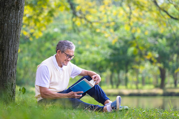 Naklejka na ściany i meble Senior Asian man reading a book while sitting under the tree by the lake at the public park for recreation, leisure and relaxation in nature concept
