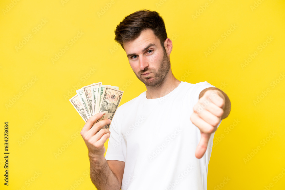 Wall mural Young caucasian man taking a lot of money isolated on yellow background showing thumb down with negative expression