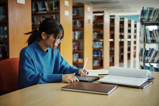Girl student with stylus leaning on hand and browsing data on tablet while sitting at table doing homework in university library.
