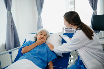 Doctor in white suit take notes while discussing and Asian elderly, woman patient who lying on bed with receiving saline solution in hospital or clinic.