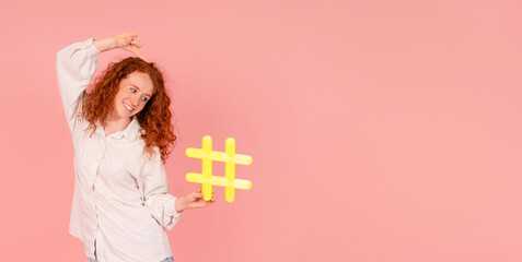 Obraz na płótnie Canvas Excited woman in white shirt holding yellow sign hashtag in her hand on pink background with copy space. social media, blogging and viral topics on internet concept