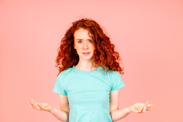 Confused red hair young woman with curly wavy hair spreading hands in shock discovering that her...
