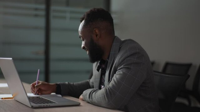 Side view portrait of smiling African-American businessman working typing keyboard on laptop computer and writing on note with pen sitting at table at office, with modern dark interior, slow motion.