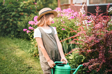 Farmer girl in summer straw hat. Little gardener farming, watering flowerbed with pink flowers, having fun in garden. Big green watering can water. Harvest help work. Cultivation plants, countryside