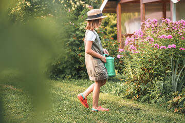 Farmer girl in summer straw hat. Little gardener farming, watering flowerbed with pink flowers, having fun in garden. Big green watering can water. Harvest help work. Cultivation plants, countryside