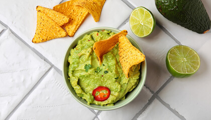 Bowl of delicious guacamole with chili pepper, nachos chips and lime on white tiled table, flat lay