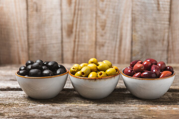 Green, black and red olives, olive oil on a brown wooden background. Fresh juicy olives in a bowl and fresh olive leaves. Vegan. Olive fruits. Place for text. Copy space.