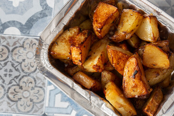 Mini roast potatoes in a takeaway foil tray carton. On a blue tile surface