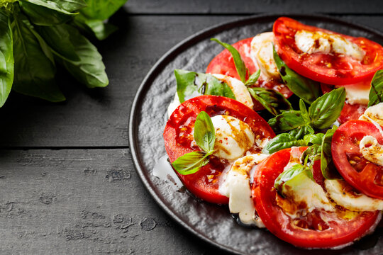 Delicious caprese salad with ripe tomatoes, mozzarella cheese and fresh basil leaves on dark wooden boards. Close up