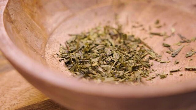 Japanese sencha dried tea leaves in rustic wooden bowl. Falling dried green ingredient in slow motion close up