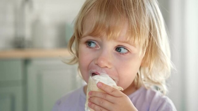 Baby girl enjoying ice cream. Pretty little toddler eating an ice-cream indoors, at home. Dining room background. Small child eats plombir and cream messy on her mouth. Cute kid with tasty sweet food.