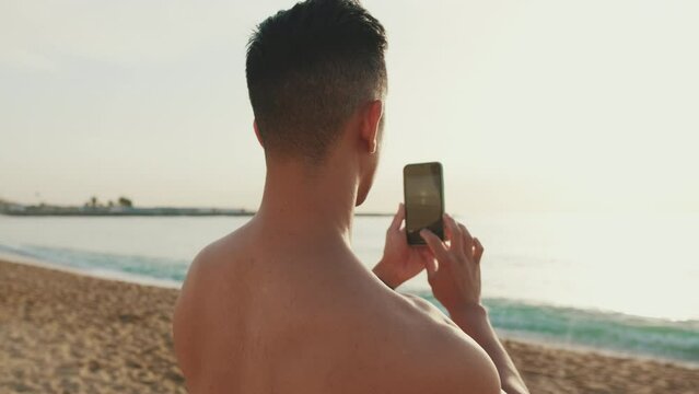 Close up, young athletic man standing on the beach filming a video on a mobile phone