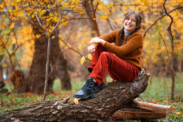 a young teenage girl posing in an autumn forest, sitting on a log and enjoying the beautiful nature and bright yellow leaves