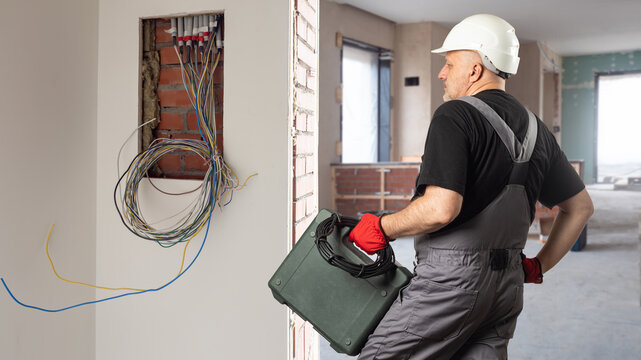 Electrician at work. Man from construction company. Place for power shield in wall. Electrician hold wire and suitcase with tools. Man in electrician uniform and hardhat. Guy from construction company