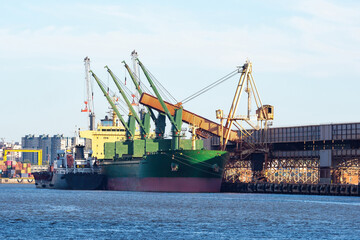 Seaport with cargo ship. Loading ship. Ship at dock in port. Merchant fleet. Sea vessel trawler. Infrastructure of commercial port. Port cranes on background blue sky. Harbor near city