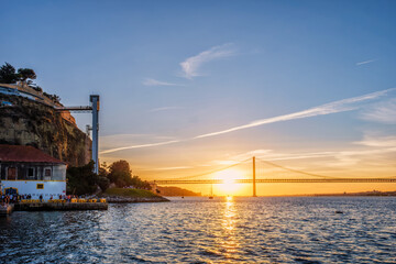 Obraz premium View of 25 de Abril Bridge famous tourist landmark of Lisbon over Tagus river and Boca do Vento Elevator and seaside restaurants with people at the sunset. Lisbon, Portugal