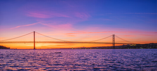 Panorama of 25 de Abril Bridge famous tourist landmark of Lisbon connecting Lisboa and Almada over Tagus river with tourist yacht silhouette at sunset. Lisbon, Portugal