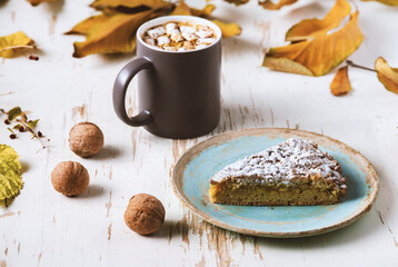 Top view of female hands holding marshmallows on shabby table with piece of cake, walnuts and scattered leaves, autumn composition