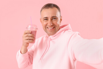 Mature man with glass of fruit smoothie taking selfie on pink background