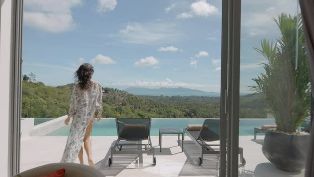Stylish white summer house interior with open terrace and swimming pool on jungle background, woman in pareo walking out to lounge area, tropical holiday concept
