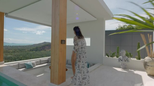Stylish white summer house terrace with open swimming pool and nature view, woman in pareo walking to coach area, tropical holiday concept