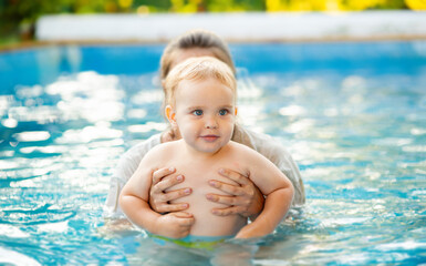 Portrait of small red-haired boy bathes in pool with hand support, baby swimming in water, summer...