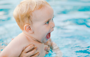 Portrait of small red-haired boy bathes in pool with hand support, baby swimming in water, summer leisure