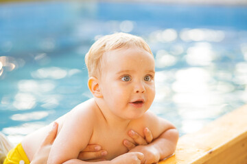 Portrait of small red-haired boy bathes in pool with hand support, baby swimming in water, summer leisure