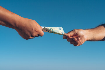 a man's hand passes 100 euros to another man's hand against the backdrop of a sea of blue sky