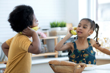 Cute little African boy pretends to use a banana to talk on the smartphone, selective focus, boy and girl playing having fun in kitchen, imagination and creativity concept