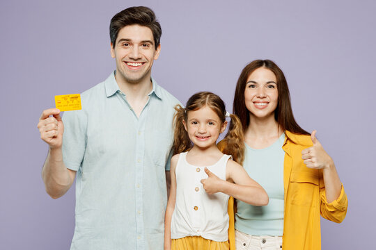 Young parents mom dad with child kid daughter girl 6 years old wearing blue yellow casual clothes hold mock up of credit bank card show thumb up isolated on plain purple background Family day concept.