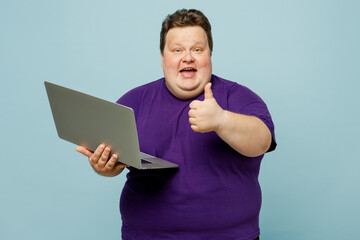 Young smiling happy fun chubby overweight IT man wears purple t-shirt casual clothes hold use laptop pc computer show thumb up isolated on plain pastel light blue cyan background. Lifestyle concept.