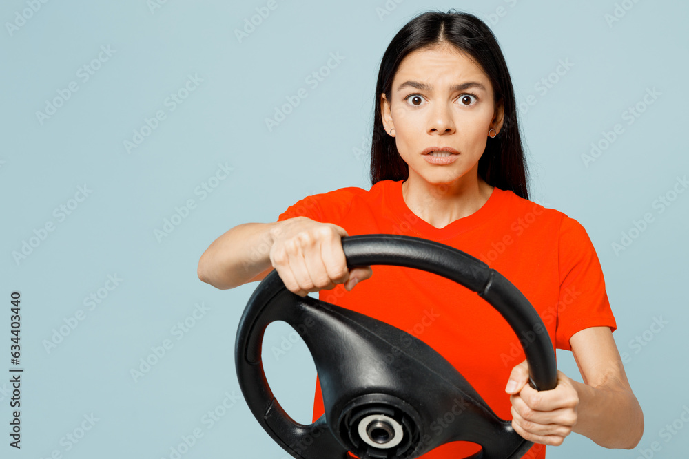 Wall mural Young shocked astonished scared sad latin woman wear orange red t-shirt casual clothes hold steering wheel driving car look camera isolated on plain pastel blue background studio. Lifestyle concept.