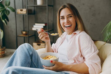 Happy young woman wear casual clothes sit in armchair eat breakfast muesli cereals with milk fruit...
