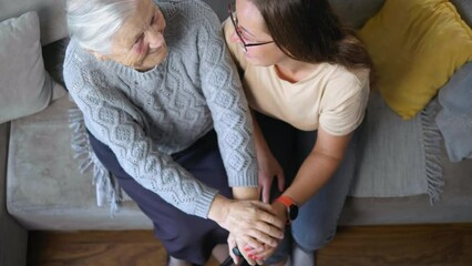 Top view shot of adult daughter hugging and holding hands of her elderly mother while sitting on sofa at home