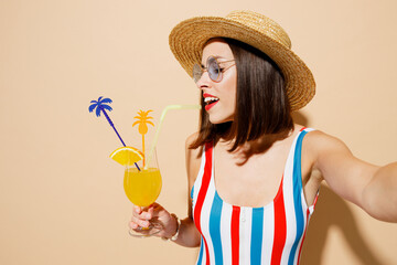 Young sexy happy woman wear one-piece swimsuit straw hat near hotel pool hold in hand orange citrus cocktail isolated on plain pastel light beige background. Summer vacation sea rest sun tan concept.
