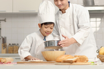 Happy Asian son and father in chef uniform with hat cooking at kitchen. cute boy child helps dad sifting flour into bowl, preparing bread dough before kneading, parent and kid in family making bakery.