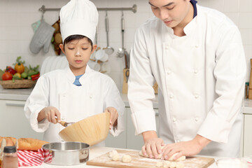 Happy Asian son and father in chef uniform with hat cook meal together at kitchen, dad parent and boy child mixing and kneading bread dough, cute family making bakery. Kid student learns to bake bread