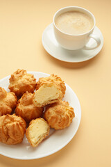 Homemade profiteroles with cream on plate with cup of coffee on beige background. Fresh baked golden profiterole. Vertical photo