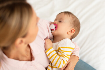Caring mother holding baby girl sleeping with pacifier, comforting kid and singing lullaby, sitting...
