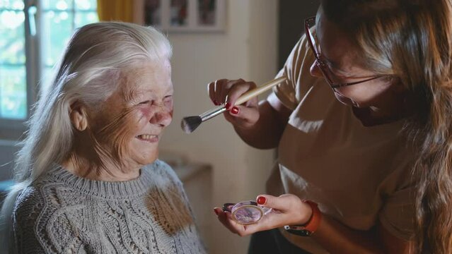 Granddaughter uses a face brush while applying make-up to her old grandmother who is uncomfortable